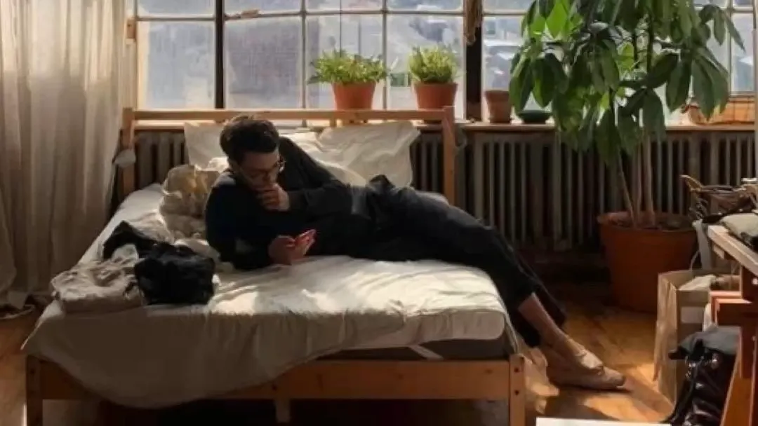A man lying on a bed in a sunlit flat, looking at his phone, a window with potted plants behind him.
