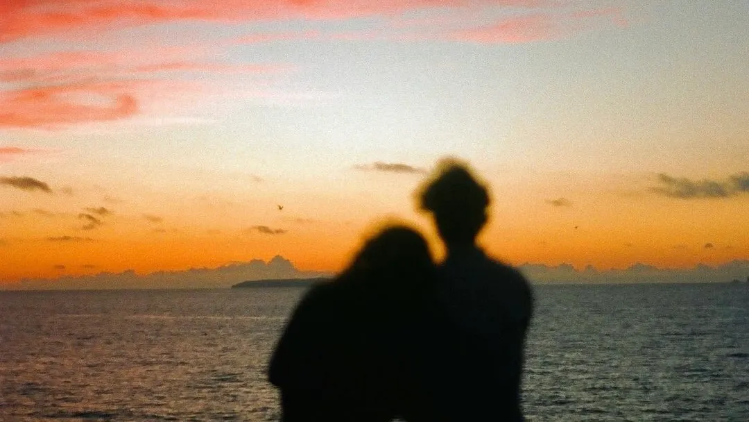 Two people silhouetted against a sunset over the sea, leaning into each other.